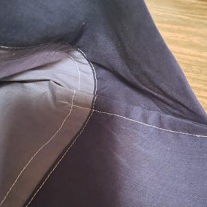 May include: A dark navy blue fabric bag featuring a lighter grey interior lining. The exterior and interior sections are joined by visible white stitching that follows the curved seam of the opening, highlighting the construction of the textile.