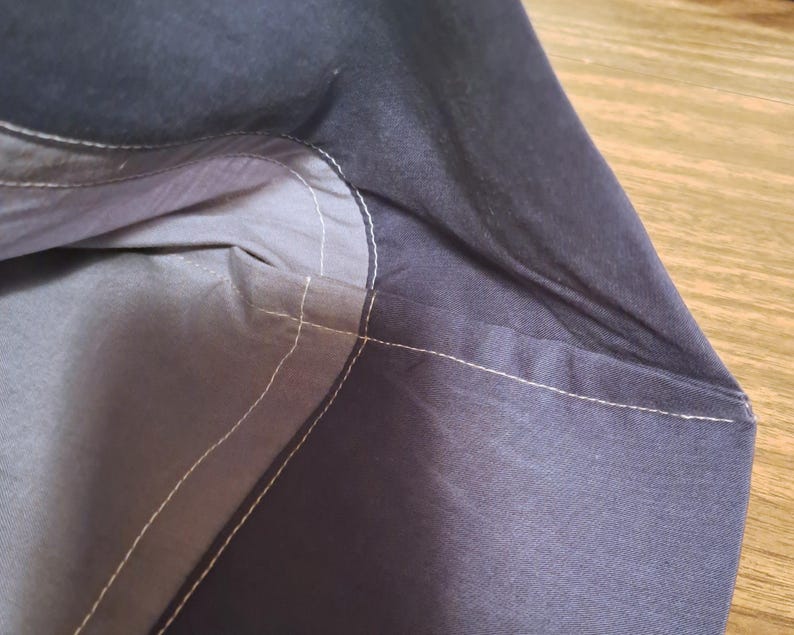 May include: A dark navy blue fabric bag featuring a lighter grey interior lining. The exterior and interior sections are joined by visible white stitching that follows the curved seam of the opening, highlighting the construction of the textile.