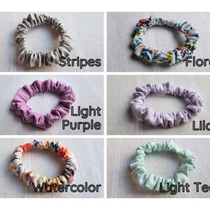 May include: A collection of six fabric hair scrunchies arranged in a grid. The scrunchies feature various designs including gray stripes, a multi-colored floral pattern, solid light purple, solid lilac, a multi-colored watercolor print, and solid light teal.