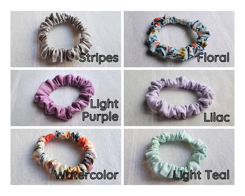 May include: A collection of six fabric hair scrunchies arranged in a grid. The scrunchies feature various designs including gray stripes, a multi-colored floral pattern, solid light purple, solid lilac, a multi-colored watercolor print, and solid light teal.