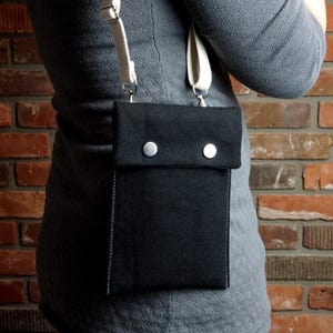 Puede incluir: Un bolso bandolera de lona negra con cierre de solapa y dos botones a presión plateados. El bolso tiene una correa blanca y se lleva sobre el hombro.