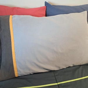 May include: A rectangular pillowcase featuring a light gray main body, a dark gray side panel, and a thin vertical orange stripe separating the two sections. The fabric appears smooth and matte, with a clean, simple design and finished edges.