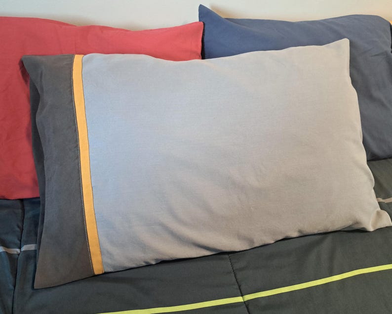 May include: A rectangular pillowcase featuring a light gray main body, a dark gray side panel, and a thin vertical orange stripe separating the two sections. The fabric appears smooth and matte, with a clean, simple design and finished edges.