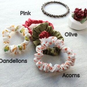 May include: Four fabric hair scrunchies in various colors and patterns. One is solid pink, one is solid olive green, one features a yellow dandelion print on white, and one features an orange acorn print on white. Text labels read Pink, Olive, Dandelions, and Acorns.