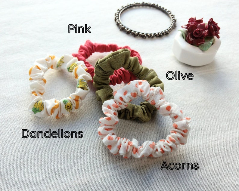 May include: Four fabric hair scrunchies in various colors and patterns. One is solid pink, one is solid olive green, one features a yellow dandelion print on white, and one features an orange acorn print on white. Text labels read Pink, Olive, Dandelions, and Acorns.