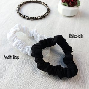 May include: Two fabric hair scrunchies, one in solid white and one in solid black, are displayed. The word White is printed near the white scrunchie, and the word Black is printed near the black scrunchie. A dark metal beaded bangle is also visible.
