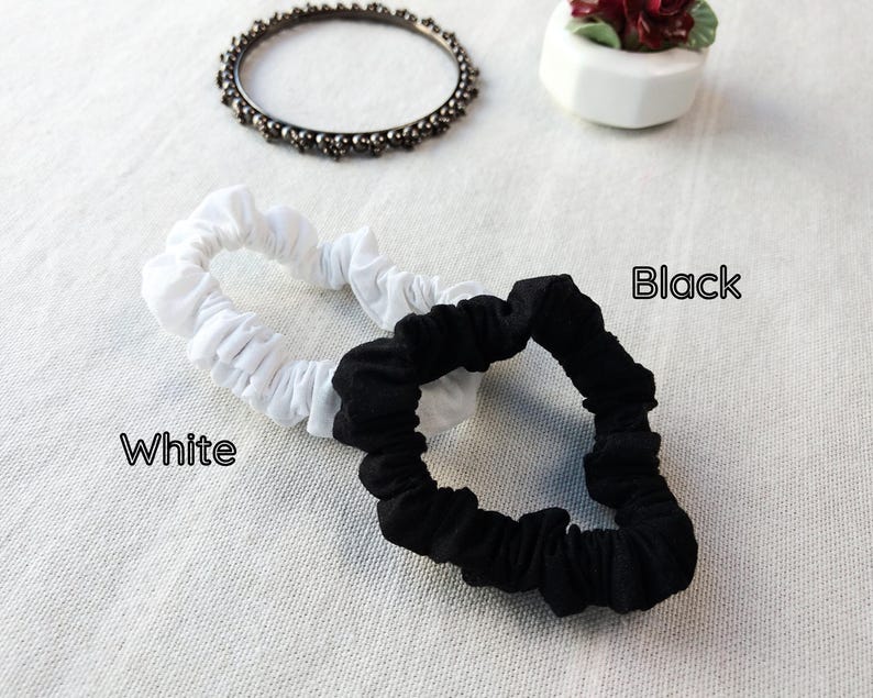 May include: Two fabric hair scrunchies, one in solid white and one in solid black, are displayed. The word White is printed near the white scrunchie, and the word Black is printed near the black scrunchie. A dark metal beaded bangle is also visible.
