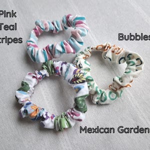 May include: Three fabric hair scrunchies. One features pink and teal stripes. Another has a white background with a multicolored floral pattern labeled Mexican Garden. The third has a white background with a brown and teal pattern labeled Bubbles.