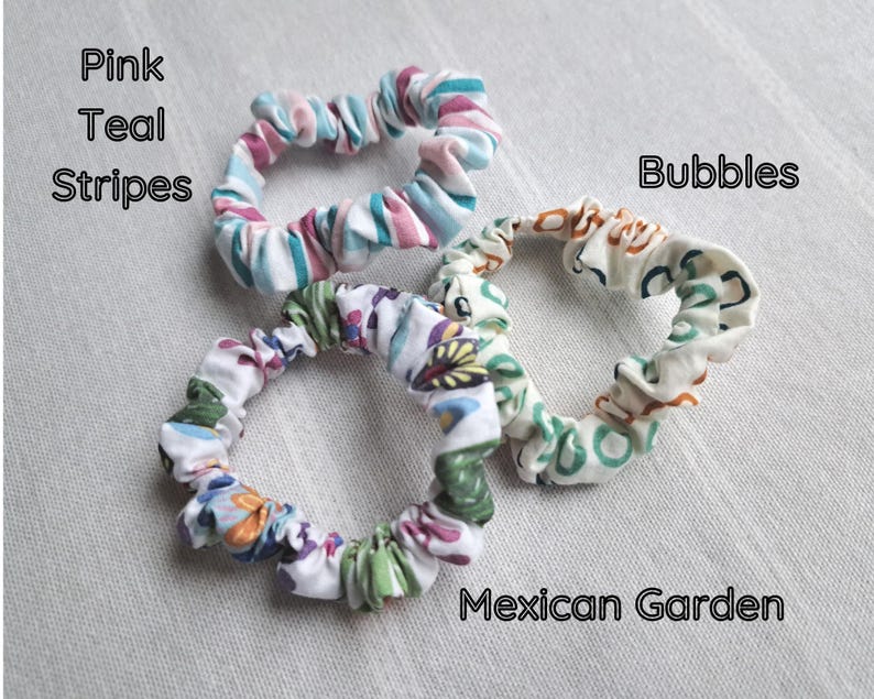 May include: Three fabric hair scrunchies. One features pink and teal stripes. Another has a white background with a multicolored floral pattern labeled Mexican Garden. The third has a white background with a brown and teal pattern labeled Bubbles.