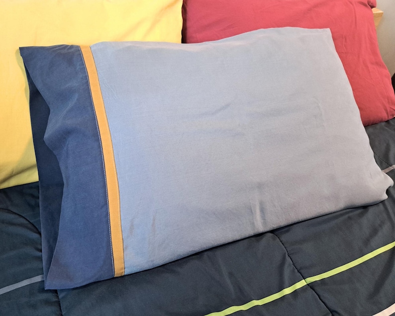 May include: A rectangular pillowcase featuring a light blue main panel, a dark blue side border, and a thin mustard yellow accent stripe separating the two sections. The fabric has a smooth, matte finish and is presented in a horizontal orientation.