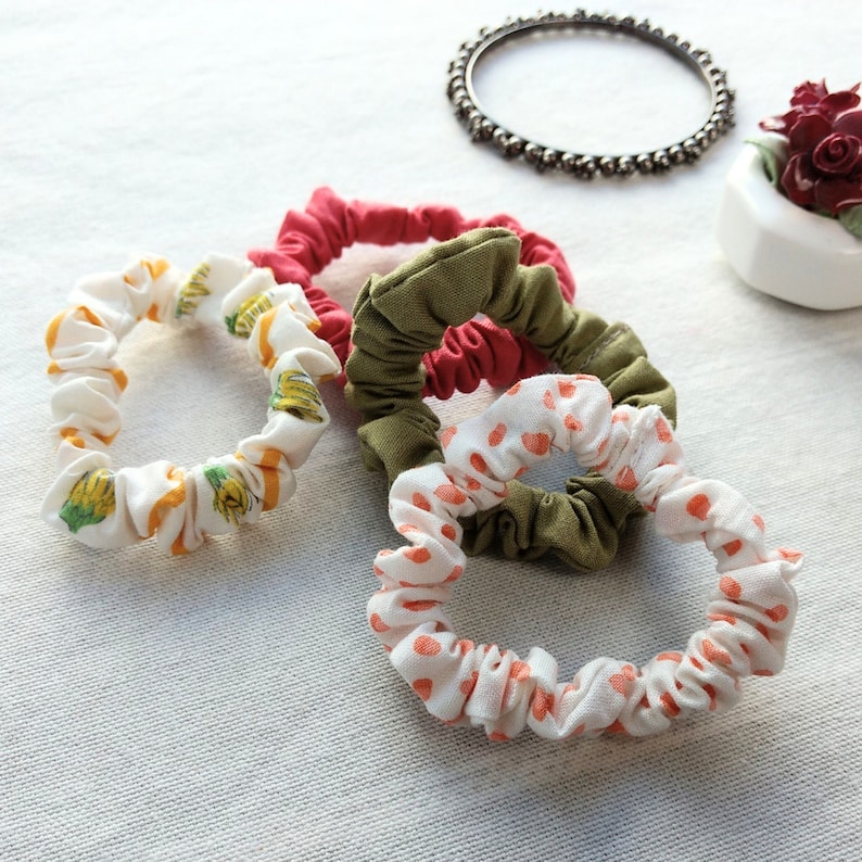4 lovely skinny scrunchies in colorful prints. There is one with pink acorns, one with yellow dandelions, one solid pink and one solid olive. In the background there is a dainty metallic bracelet and a porcelain floral decoration.