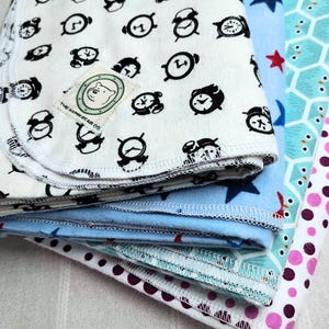 May include: A stack of four folded fabric cloths with finished serged edges. The top cloth is off-white with a black alarm clock pattern and a square label reading THE HAPPY BEAR CO. Below are blue fabric with stars, teal with a geometric pattern, and white with purple polka dots.