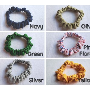 May include: A collection of six fabric hair scrunchies in various colors and patterns. The set includes solid navy, olive, green, silver, and yellow scrunchies, along with one pink floral patterned scrunchie. Each item is labeled with its color name.