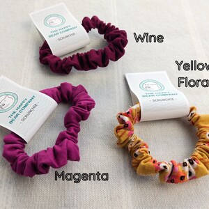 May include: Three fabric hair scrunchies with white product tags labeled The Happy Bear Company. One is wine-colored, one is magenta, and one features a yellow floral pattern. The text Wine, Magenta, and Yellow Floral identifies each item.