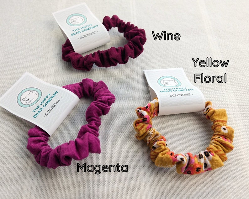 May include: Three fabric hair scrunchies with white product tags labeled The Happy Bear Company. One is wine-colored, one is magenta, and one features a yellow floral pattern. The text Wine, Magenta, and Yellow Floral identifies each item.