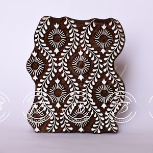 Puede incluir: Un bloque de madera marrón oscuro con un diseño floral repetitivo en blanco. El patrón consta de formas circulares y de hojas. Este bloque probablemente se utiliza para imprimir o estampar.