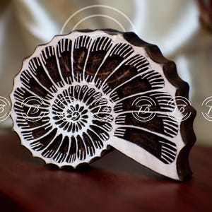 May include: A wooden, half-shell shaped carving with a detailed spiral pattern. The design features dark brown lines against a white background, creating a contrasting effect. The object is placed on a dark wooden surface.