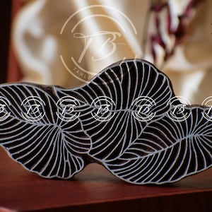 May include: A dark brown wooden block with a leaf design. The block features a detailed pattern of white lines that create the veins of the leaves. The background is a blurred mix of cream and burgundy.