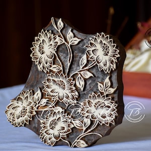 Peut inclure: Un bloc d'impression en bois brun foncé présentant un motif floral sculpté de cinq fleurs ressemblant à des chrysanthèmes avec des tiges et des feuilles. Les surfaces en bois clair en relief créent un contraste avec le fond sombre.