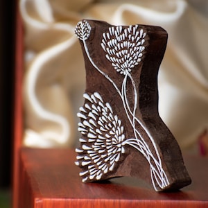 Puede incluir: Un bloque de madera marrón oscuro con un diseño floral tallado. El diseño presenta flores y tallos blancos estilizados. El bloque se muestra sobre una superficie marrón rojizo, con una tela suave de color crema en el fondo.
