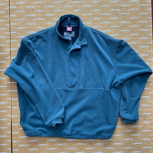 May include: A teal fleece pullover with a stand-up collar and a snap closure. The long-sleeved top features a front pocket and the brand name "Simms" embroidered on the chest. The garment is made of a soft, warm material.