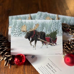 May include: A Christmas card featuring a painting of a moose pulling a red sled with a Christmas tree through a snowy forest. The card has the artist's name and location printed on it, along with a copyright notice.