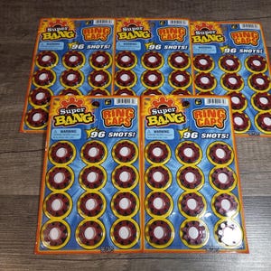 Op de afbeelding: Vijf pakketten Super Bang Ring Caps, elk met 96 schoten. De pakketten zijn geel en blauw met rode en witte ringkappen die zichtbaar zijn in de verpakking.