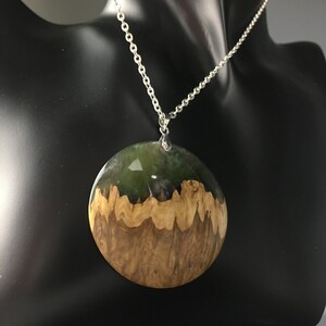 May include: A round pendant necklace with a silver chain. The pendant is made of wood and resin, with a green and brown color scheme.