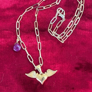 Puede incluir: Un collar de cadena dorada con un colgante de corazón alado y un dije de piedra preciosa púrpura. El collar está sobre un fondo de terciopelo rojo.