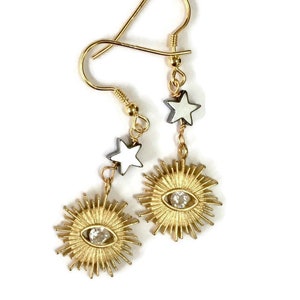 Puede incluir: Pendientes de oro con un diseño de sol y un ojo de cristal en el centro. Cada pendiente tiene dos pequeñas estrellas blancas colgando de él.