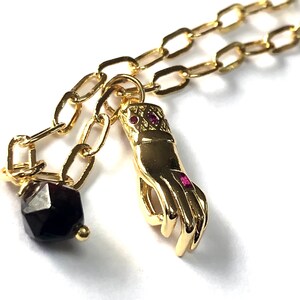 May include: A gold chain necklace with a hand charm and a dark red gemstone pendant.
