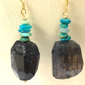Op de afbeelding: Gouden oorbellen met een grote, gefacetteerde, donkergrijze steen en een stapel turquoise en aquablauwe kralen.