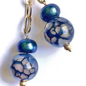 Puede incluir: Un par de pendientes colgantes con una cadena dorada, una perla azul iridiscente y una cuenta de ágata azul con un patrón blanco.