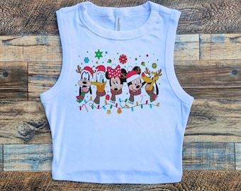 Könnte beinhalten: Weißes ärmelloses Crop-Top mit einem weihnachtlichen Druck, der Mickey Mouse, Minnie Mouse, Donald Duck, Goofy und Pluto zeigt, die Weihnachtsmützen und Schals tragen. Der Druck enthält eine Lichterkette und Zuckerstangen.
