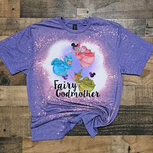 May include: Purple t-shirt with a bleach-splattered design featuring three fairies from the Disney movie Cinderella. The fairies are flying with wands and the text "Fairy Godmother" is printed below them.