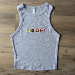 May include: A light grey sleeveless tank top featuring a row of small embroidered icons across the chest, including a yellow star, a black bomb, a colourful flower, and a red and white mushroom. The garment has a ribbed crew neckline.