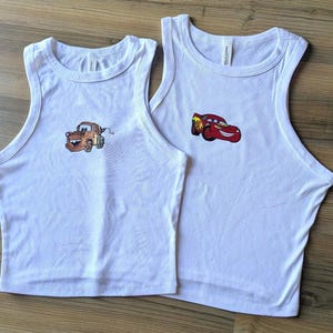 May include: Two white sleeveless tank tops featuring embroidered character designs. The left tank top displays a brown tow truck character, while the right tank top features a red race car character with yellow and orange lightning bolt graphics.