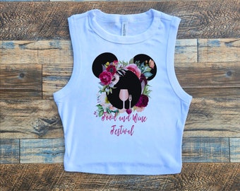 May include: White crop top with a floral design in the shape of Mickey Mouse ears. The design includes a wine glass and fork, with the words "Food and Wine Festival" in a cursive font.