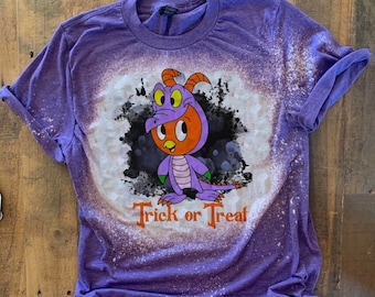 May include: A purple t-shirt with a distressed bleach effect. The shirt features a cartoon dragon in an orange and purple costume, with the words "Trick or Treat" in orange text below the dragon. The shirt is on a wooden surface.