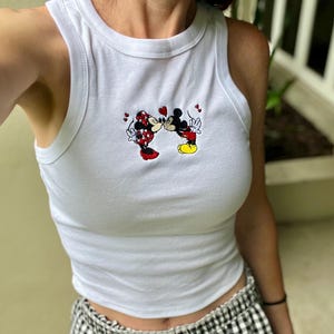 May include: White tank top with a crew neck featuring an embroidered image of Mickey and Minnie Mouse kissing. The tank top is paired with black and white checkered shorts.