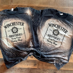 K&ouml;nnte beinhalten: Zwei schwarze T-Shirts mit einem Design aus Bleichmittelspritzern. Die Shirts zeigen ein wei&szlig;es Etikett mit dem Text "Winchester Brothers Since 1983 Saving People Hunting Things A Family Business" und ein Pentagramm-Symbol.