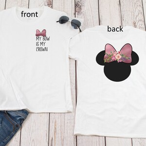 May include: White t-shirt with the text "My Bow Is My Crown" and a pink bow on the front. The back of the shirt features a black silhouette of Mickey Mouse ears with a pink bow and floral embellishments.