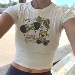 May include: Cream-colored crop top with a graphic featuring Mickey Mouse ears, a coffee cup, sunglasses, and a park ticket. The text "literally just a girl" is above the graphic.