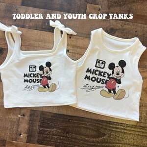 May include: Two white crop tank tops with a Mickey Mouse graphic and the text "Mickey Mouse" printed on the front. The tops are made of a soft, stretchy material and have a relaxed fit.