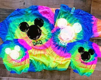 May include: Four tie-dye t-shirts with a Mickey Mouse silhouette design. The shirts are in various colors, including pink, blue, green, and yellow. The shirts are labeled "Dad", "Mom", "Teddy", and "Sissy".