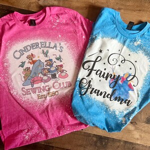 May include: Two bleach-splattered t-shirts with Disney-themed designs. The pink shirt features Cinderella's mice and the text "Cinderella's Sewing Club Est. 1950". The blue shirt features a fairy godmother and the text "Fairy Grandma".