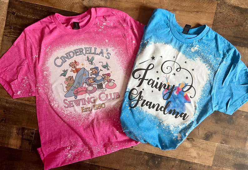 May include: Two bleach-splattered t-shirts with Disney-themed designs. The pink shirt features Cinderella's mice and the text "Cinderella's Sewing Club Est. 1950". The blue shirt features a fairy godmother and the text "Fairy Grandma".