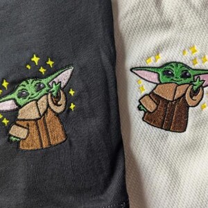 May include: A black fabric and a white textured fabric, each featuring an embroidered design of a small green creature with large ears wearing a brown robe. The creature is depicted with one hand raised and surrounded by small yellow embroidered stars.