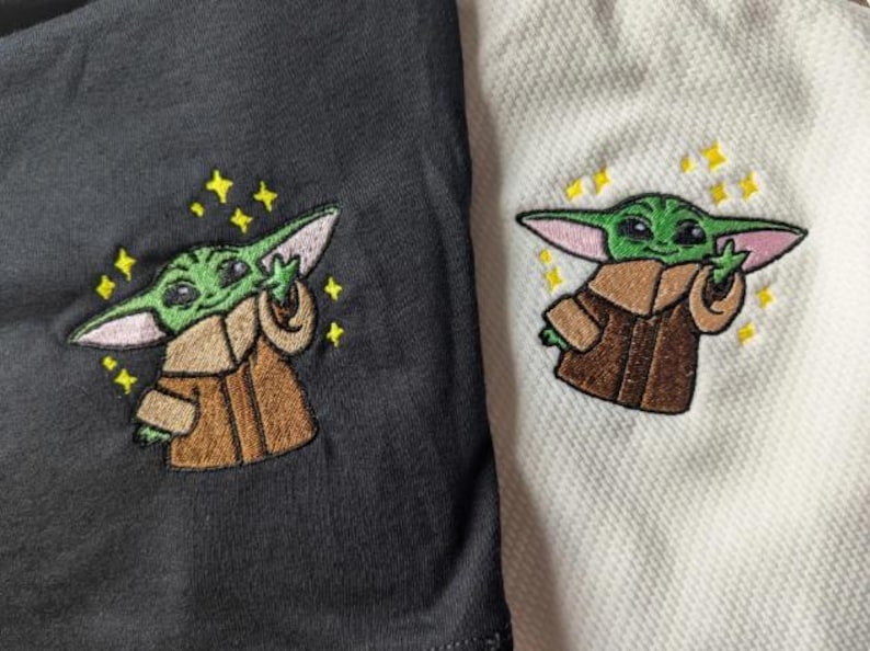 May include: A black fabric and a white textured fabric, each featuring an embroidered design of a small green creature with large ears wearing a brown robe. The creature is depicted with one hand raised and surrounded by small yellow embroidered stars.