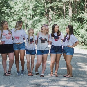 May include: Six friends wearing white t-shirts with a black and white Mickey Mouse head design and floral accents. They are standing on a dirt road with trees in the background.